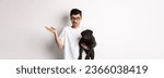 Small photo of Image of handsome young man holding black pug and looking confused. Guy shrugging shoulders and staring indecisive at camera, carry dog in arm, white background.