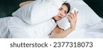 Small photo of Portrait of asian woman waking up in bed, looking shocked at mobile phone, realise she overslept.