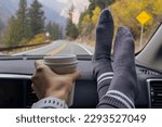 Woman on a passenger seat holding her feet and a cup of coffee on the dash, while travelling throught the fall mountains