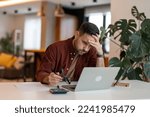 Small photo of Man working on laptop while sitting at his working place in his apartment. Tired brown haired man taking glasses off working too long at computer. Exhausted male suffer from headache.