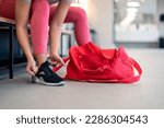 Small photo of Cropped shot of fit sports woman in sportswear with gym bag wearing pink yoga pants and sneakers sitting on bench, getting ready for exercise session, tying her shoelaces in locker room at gym.