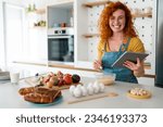 Small photo of Joyous smiling young adult redhead woman looking at camera while searching food recipes on digital tablet in the kitchen, watching an online cooking class .