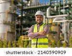 Small photo of Asian engineer man with white safety helmet standing front of oil refinery. Industry zone gas petrochemical. Factory oil storage tank and pipeline. Workers work in the refinery construction building