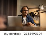 Small photo of Young asian man host streaming podcast with condenser microphone work on laptop at small broadcast home studio. Content creator blogger recording voice over radio interview guest conversation