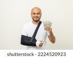 Small photo of Happy young asian man broken arm holding Dollar banknote. Man put on plaster bandage cast splint. Male patient wearing sling support arm. life insurance and accident have money to spend while sick