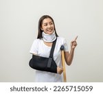 Small photo of Happy young asian woman broken arm pointing finger on isolated background. Female put on plaster bandage cast splint. Patient wearing sling arm after accident injury. life insurance and accident