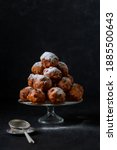 Small photo of A pile of traditional oliebollen (Dutch dough firtters) on a glass stand on black background with small sieve next to it.