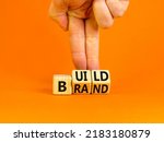 Small photo of Build your brand symbol. Concept words Build brand on wooden cubes. Businessman hand. Beautiful orange table orange background. Build your brand and business concept. Copy space.