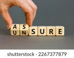 Small photo of Assure or unsure symbol. Businessman turns wooden cubes and changes the concept word unsure to assure. Beautiful grey table, grey background, copy space. Business and assure or unsure concept.