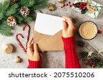 Desktop of woman hands in red sweater holding blank paper and envelope . Flat lay of gray background with cup of coffee and Christmas decoration. Top view mock up and copy space for text.