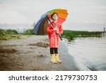 Small photo of Boy in a red raincoat and yellow rubber boots stands at river bank and holding rainbow umbrella. School kid standing still near autumn lake. Child wearing waterproof clothes at shoreside.