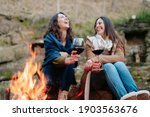 young happy women laughing, holding glass of red wine. Females warming next to the fire. Campfire, outdoors activities concept.