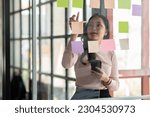 Small photo of Asian businesswoman using mobile phone with pen to write on notepad business plan idea and find flaws to prevent mistakes in work.