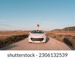white European girl sits in the seat of her white convertible car and raises her hands to the sky as she rides on a summer afternoon.