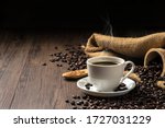 Hot coffee in a white coffee cup and many coffee beans placed around and sugar on a wooden table in a warm, light atmosphere, on dark background, with copy space.