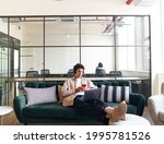 Relaxed casually dressed young man with mobile phone and wireless headphones working on laptop in seating area of modern open plan office
