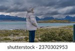 Small photo of A man in a down jacket and a hat is standing on an island in the Beagle Channel, looking into the distance. Profile view. There is scant stunted vegetation on stony soil. A picturesque mountain range