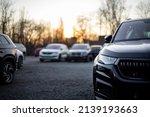 Partial front view of new black modern family SUV. Visible front LED light and part of gloss black grille and bumper. Out-of-focus cars and trees in the background. Sunset light. Parking on gravel.