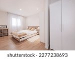Small photo of Bedroom with wooden headboard, white hanging tables, beige cushions and matching blanket, wooden bookcase and window with white curtains and white cabinets in one corner