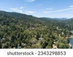Small photo of aerial shot of majestic mountain ranges covered with tall lush green trees and homes along the hillside with a lake, blue sky and clouds at Lake Gregory in Crestline California USA