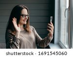 Happy brunette woman in glasses making facetime video calling with smartphone at home, using zoom meeting online app, social distancing, work from home, work remotely concept
