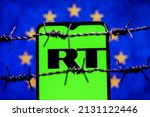 Small photo of Kazan, Russia - Mar 02, 2022: Smartphone with RT (Russia Today) logo on background of flag of European Union behind barbed wire. Russia Today network banning in EU.