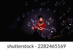 Small photo of Magician lady make soap bubbles show, an illusionist in theatrical clothes posing at black background. Woman actress in theatrical costume. Concept of stage perform and fun show. Copy ad text space
