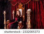 Small photo of Lovely lady in an old style on antique throne fortress, looking at camera. Medieval queen in historical image posing sitting on golden throne in castle room. Concept of theatrical. Copy ad text space