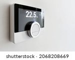 Screen of a smart digital thermostat with the external and target temperatures indicated. Rotary dial to control and adjust. Scheduling and programmable domestic heating. White wall and black frame