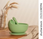 Small photo of Modern green silicone bowl with suction base and spoon on wooden table near glass vase with dried flowers. Serving food, baby tableware, first feeding concept. Instagram use, square frame, soft focus.