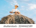 Small photo of Handsome school aged boy sitting at top of haystack play ukulele. Carefree child resting on hayrick, from below view. Outdoor walking. Beautiful blue sky. Summer vacation, countryside lifestyle.