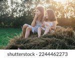 Small photo of Pretty little girls sitting together at top of haystack on sunny day. Carefree children resting on hayrick. Outdoor walking. Happy sisters. Summer vacation, countryside lifestyle, haying.