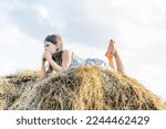 Small photo of Thoughtful little girl lying on stomach at top of haystack. From below portrait, side view. Child resting on hayrick. Outdoor walking. Beautiful blue sky. Summer vacation, countryside lifestyle.
