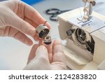 Small photo of Female hands hold a metal bobbin case and bobbin on the background of a sewing machine. Close-up of hands. Tailor workflow details.