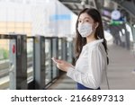 Young Asian professional business woman stands on a sky train station in town wearing a face mask while uses her smartphone to send messages  to the customer office