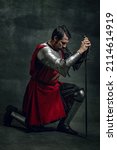 Small photo of Taking an oath. Vintage portrait of brutal seriuos man, medieval warrior or knight with dirty wounded face holding sword isolated over dark background. Comparison of eras, history, renaissance style