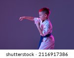 Small photo of Pupil. Portrait of kid, male taekwondo, karate athletes in doboks doing basic movements isolated on purple background in neon. Concept of sport, education, skills, martial arts, healthy lifestyle.