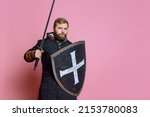 Portrait of young brave man, medieval warrior or archer with sword and protective shield isolated on pink studio background. Looks seriour and fearless. Comparison of eras, history, renaissance style.
