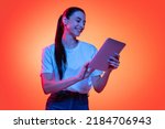 Remote studying. Portrait of young emotional girl, student in white t-shirt isolated on orange color background in neon light. Concept of beauty, art, fashion, education