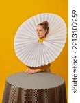 Small photo of Dinner party. Young indifferent girl in giant jabot collar or neckwear and yellow tights isolated over yellow background. Contemporary art, weird beauty, avant-garde fashion.