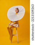 Small photo of Young indifferent girl in giant jabot collar or neckwear and yellow tights isolated over yellow background. Contemporary art, weird beauty, avant-garde fashion.