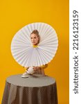 Small photo of Tea party. Young indifferent girl in giant jabot collar or neckwear and yellow tights isolated over yellow background. Contemporary art.
