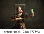 Portrait of pretty young girl, princess with bubble gum, holding picture frame and taking selfie with phone against dark green background. Concept of history, renaissance art, comparison of eras