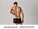 Small photo of Back pains. Muscular man standing in underwear and holding his back against grey studio background. Medical treatment. Concept of men's health and beauty, body care, fitness, wellness, ad
