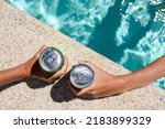 Two hands hold cans of cold drink in the pool on a hot day. The concept of vacation in a friendly company. Top view, selective focus on the bank.