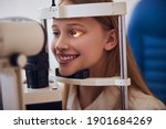 Small photo of Smiling blonde Caucasian female child in blue medicine chair and looking to slit lamp machine during medical check up in eyes clime
