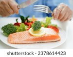 Small photo of Asian elderly woman patient eating salmon stake and vegetable salad for healthy food in hospital.