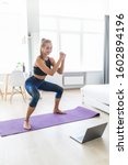 Small photo of Young attractive woman in sportswear making deep sumo squats in living room while watching fitness online video on laptop. Lady doing exercises for maintaining optimum weight and good physical shape