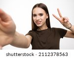 Image of pleased brunette woman laughing and showing peace sign while taking selfie photo on cellphone isolated over white background