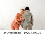 A girl in a peach warm fleece tracksuit smiles with her eyes closed and lies on the shoulder of another girl in a gray fleece suit against a background of white brickwork with space for text.